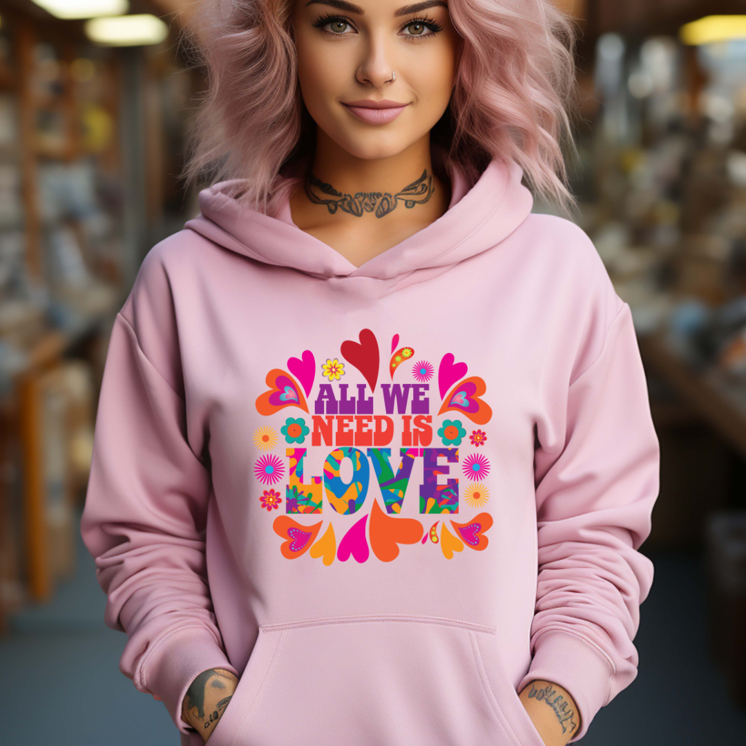 Young woman with pink hair wearing a pink All We Need Is Love hoodie with colorful hippie flowers and hearts created by graphic apparel shop Midnight Gypsy Designs as part of their slogan shirt collection. Perfect mental health awareness shirt, Vintage 1960s hippie hoodie, positive vibes hoodie, all we need is love shirt, cute hippie shirt, vintage 1960s shirt, mental health awareness hoodie, cute love shirt, valentine's day shirt, valentine's day hoodie, cute kindness hoodie, and cute kindness shirt.
