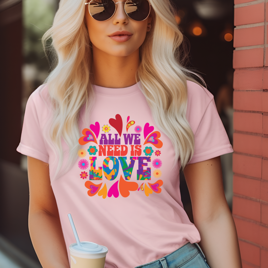 Young woman with blonde hair wearing sunglasses and holding a coffee cup while wearing a pink All We Need Is Love T-shirt by graphic apparel company Midnight Gypsy Designs. Perfect All We Need is Love shirt, mental health awareness shirt, 60s love shirt, cute hippie shirt, flower child shirt, colorful peace shirt, and cute kindness shirt.