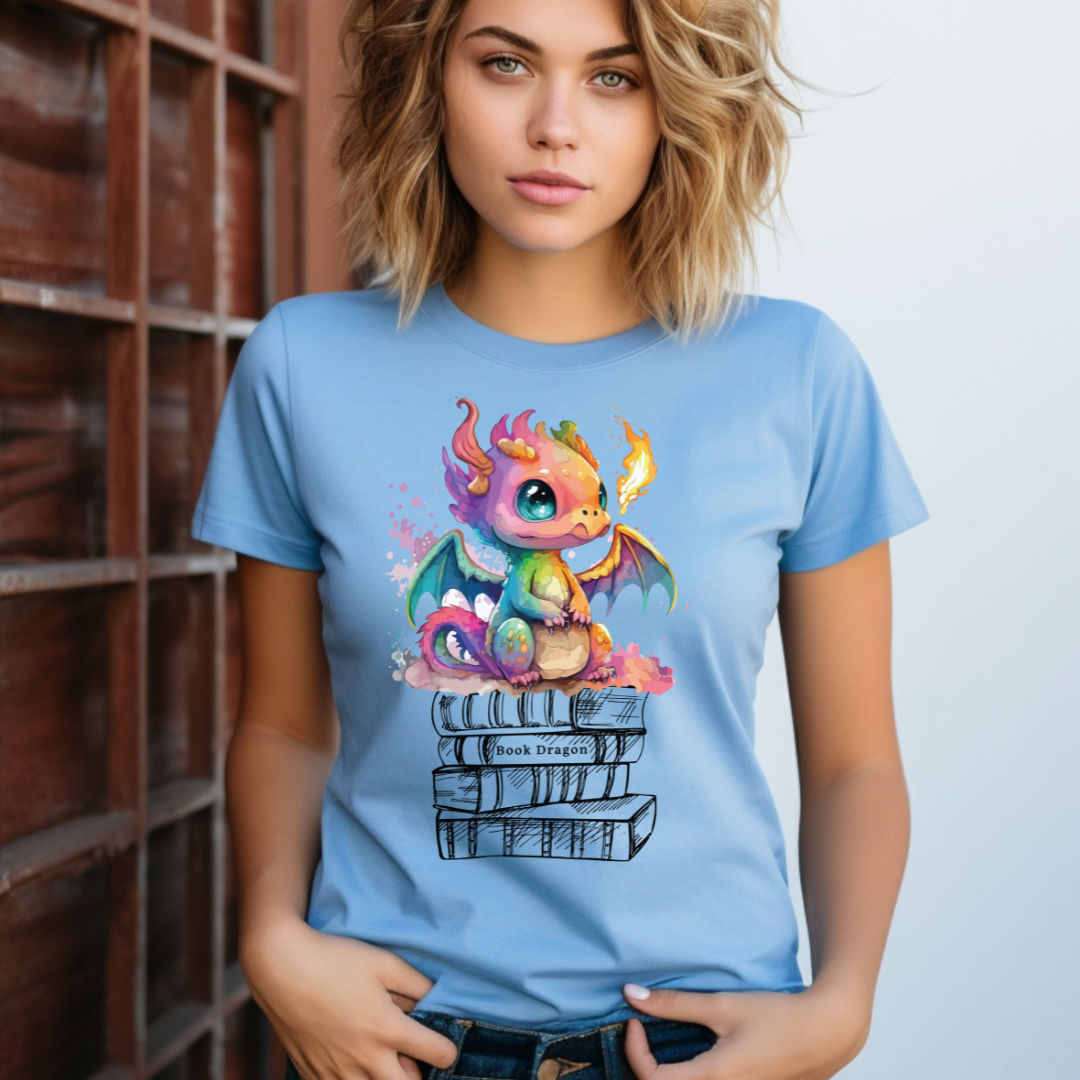 Young woman with shoulder length blonde hair wearing blue book dragon shirt with a fire breathing rainbow dragon sitting on top of a stack of books designed by graphic apparel shop Midnight Gypsy Designs as part of their bookish merch collection. Cute dragon shirt for fantasy readers!