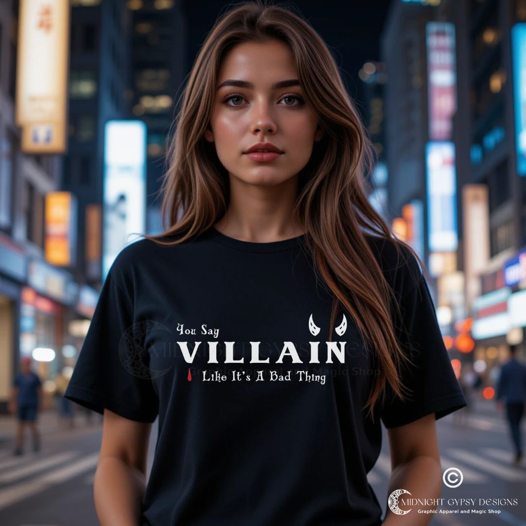 Young woman with long brown hair in the city with bright city lights wearing a black Villain shirt that says You Say Villain Like It's A Bad Thing created by graphic apparel company Midnight Gypsy Designs. Cute villain shirt, dark fantasy shirt, villain book shirt, villain bookish shirt, villain lover shirt, villain lover t-shirt, villain bookish t-shirt, cute Halloween shirt, devil villain tee, villain book merch, anti-hero bookish shirt, Disney villain shirt, and dark fantasy book shirt.