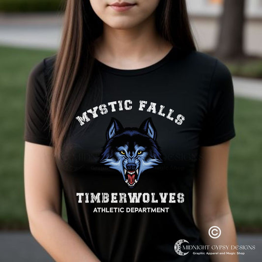 Young woman with long brown hair standing in Mystic Falls wearing a black Mystic Falls Timberwolves shirt with a blue werewolf showing its fangs created by graphic apparel company Midnight Gypsy Designs as part their The Vampire Diaries merch collection. Perfect The Vampire Diaries shirt, Mystic Falls shirt, Mystic Falls Timberwolves t-shirt, Stefan Salvatore shirt, Stefan Salvatore t-shirt, Stefan Salvatore tee, Stefan Salvatore 17 shirt, Mystic Falls High School shirt, Team Stefan shirt, Team Damon Shirt.
