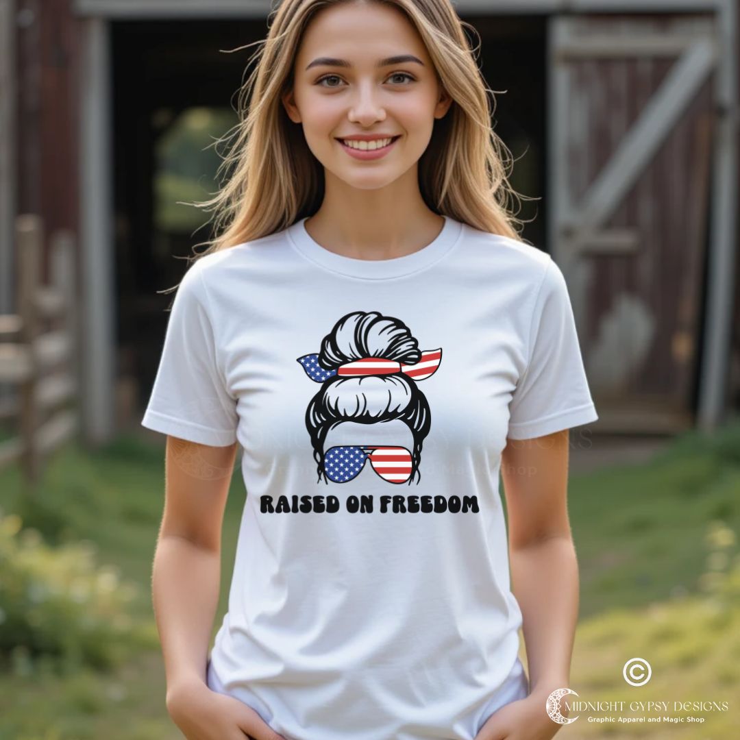 Young woman standing in front of a barn wearing a white Raised on Freedom shirt with graphic of a woman with an American flag hair tie and sunglasses created by graphic apparel company Midnight Gypsy Designs as part of their patriotic apparel collection. Perfect USA patriotic shirt, All American Girl shirt, girl power shirt, American pride shirt, USA shirt, USA flag shirt, womens patriotic shirt, Fourth of July shirt, Patriotic pride shirt, American flag shirt, Independence Day shirt, & American girl shirt.