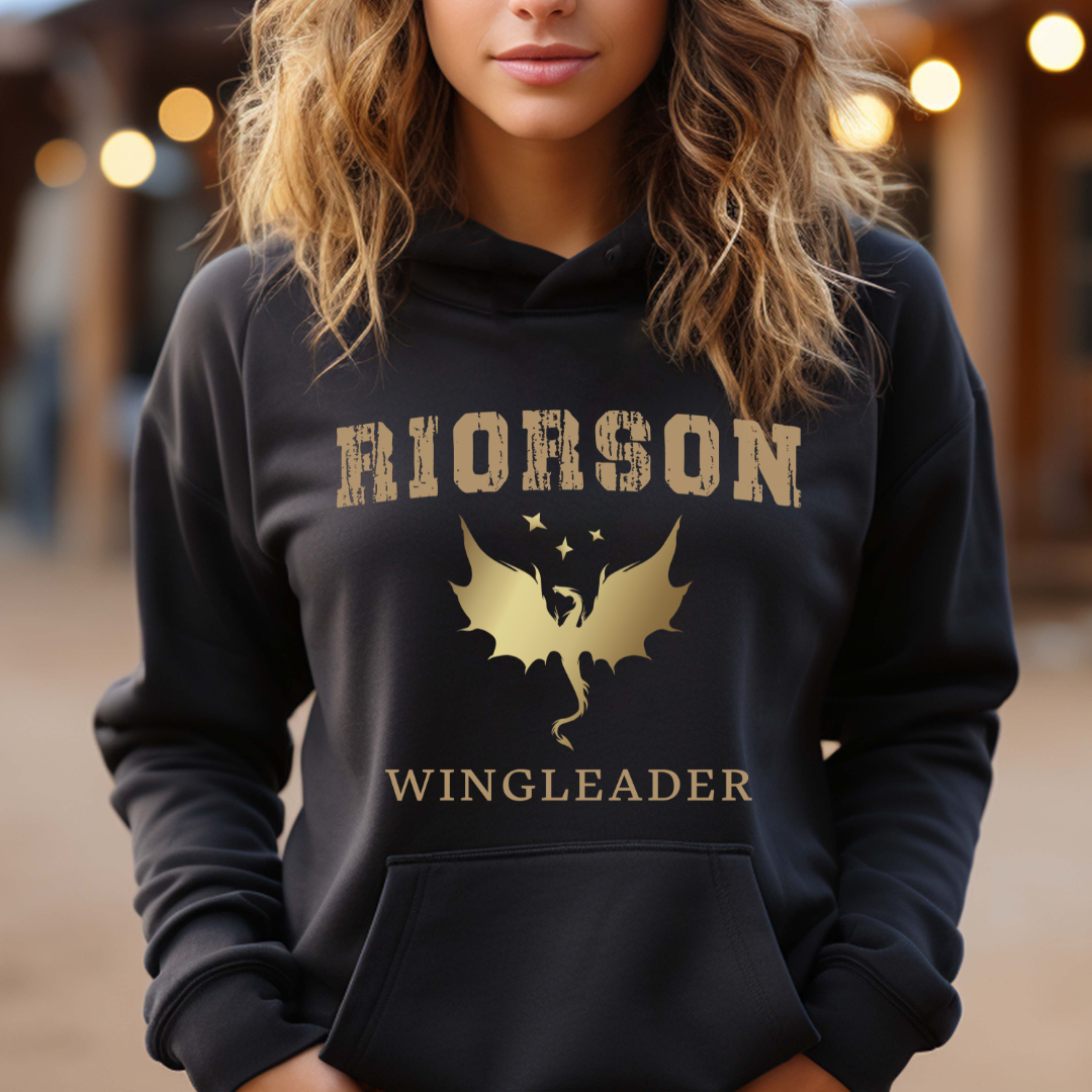 Young woman with wavy blonde hair standing outside wearing a black Fourth Wing shirt that says Riorson Wingleader with a dragon and stars created by graphic apparel company Midnight Gypsy Designs as part of its bookish merch collection for fantasy book readers. Perfect dragon riders shirt, Xaden Riorson shirt, Iron Flame shirt and Onyx Storm shirt.