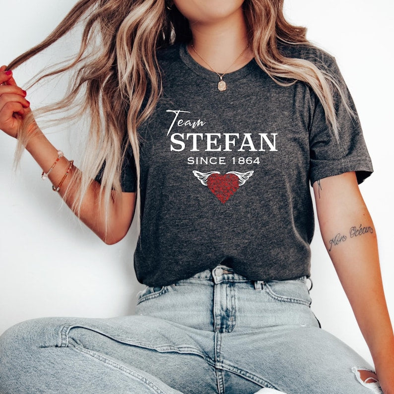 Young woman with long blonde hair wearing denim jeans and a dark grey heather t-shirt that reads The Vampire Diaries Team Stefan Since 1864 with a red heart created by bookish shop Midnight Gypsy Designs.
