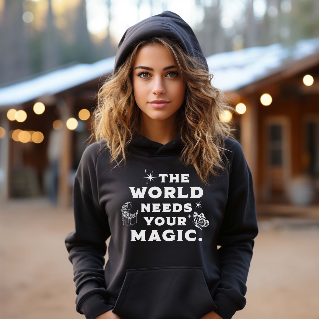 Young woman with long hair wearing a black The World Needs Your Magic hoodie with the moon and stars and butterfly created by graphic apparel company Midnight Gypsy Designs for their kindness apparel collection. Perfect mental health awareness shirt and cute kindness shirt.