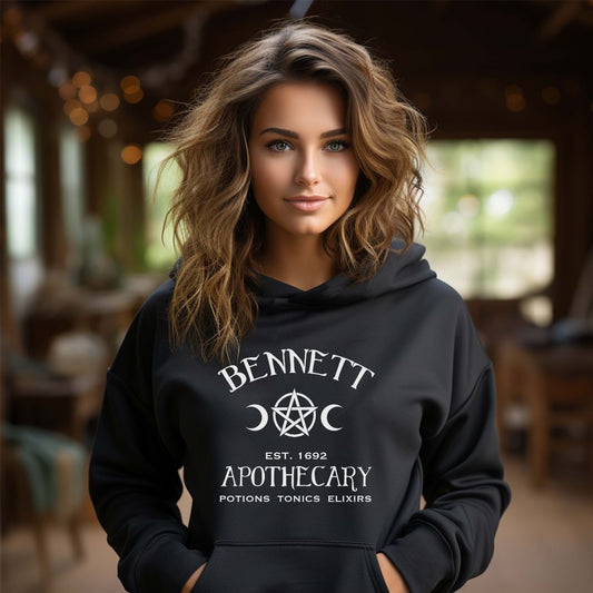 Young woman with long light brown hair wearing a Gildan black hoodie with white lettering that reads Bennett Apothecary Established 1692 Potions Tonics Elixirs inspired by Bonnie Bennett from The Vampire Diaries and created by apparel apparel company Midnight Gypsy Designs. Perfect The Vampire Diaries hoodie, Bonnie Bennett hoodie, Bonnie Bennett shirt, Salem witch trials shirt, Mystic Falls hoodie, Vampire Diaries shirt, Mystic Falls shirt, Bonnie Bennett Apothecary shirt, and Salem witch trials hoodie.