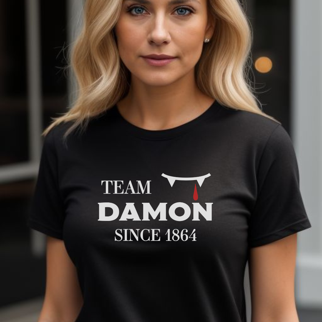 Young woman with long blonde hair wearing a black The Vampire Diaries Team Damon Since 1864 shirt with fangs and blood drip created by graphic apparel company Midnight Gypsy Designs. Perfect Team Damon shirt, Damon Salvatore shirt, the Vampire Diaries shirt, cute vampire shirt, team Damon apparel, Salvatore Brothers shirt, team Damon t-shirt, Mystic Falls shirt, and Mystic Falls t-shirt for fans of The Vampire Diaries!