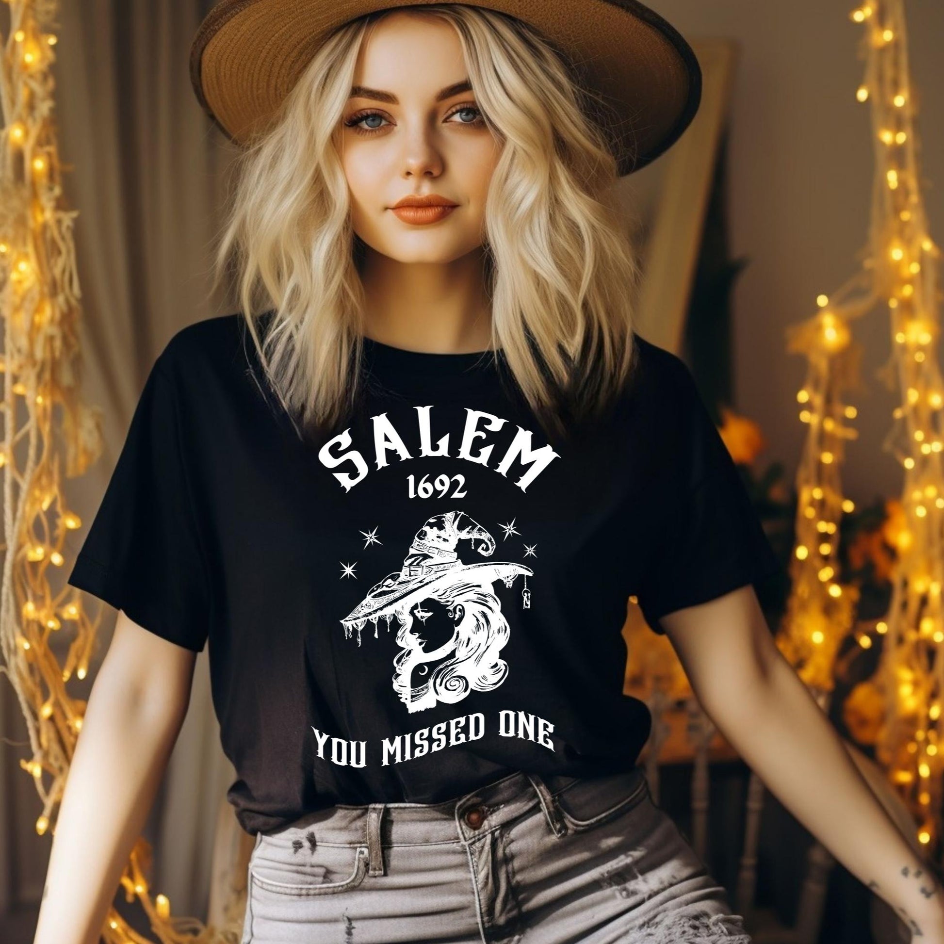 Young woman with blonde hair and blue eyes wearing grey pants and a black t-shirt with 1692 Salem Witch Trials You Missed One written in white letters and a witch in a witch hat from apparel shop Midnight Gypsy Designs.