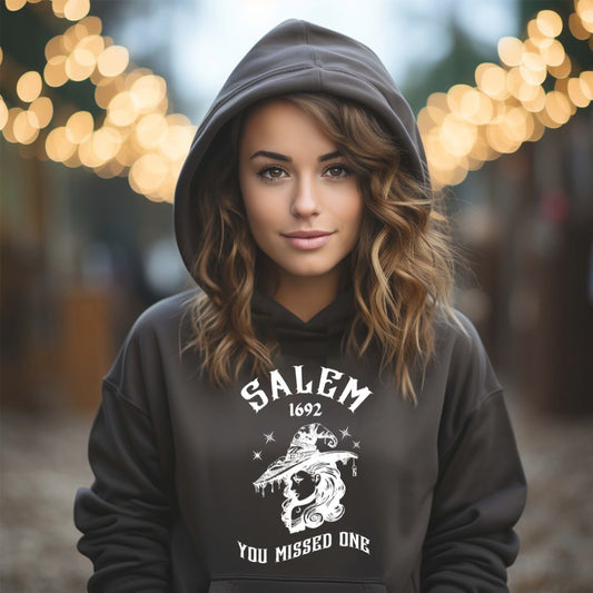 Young woman with long brown hair wearing black Gildan hoodie with 1692 Salem Witch Trials You Missed One written in white letters and a witch in a witch hat from apparel shop Midnight Gypsy Designs.