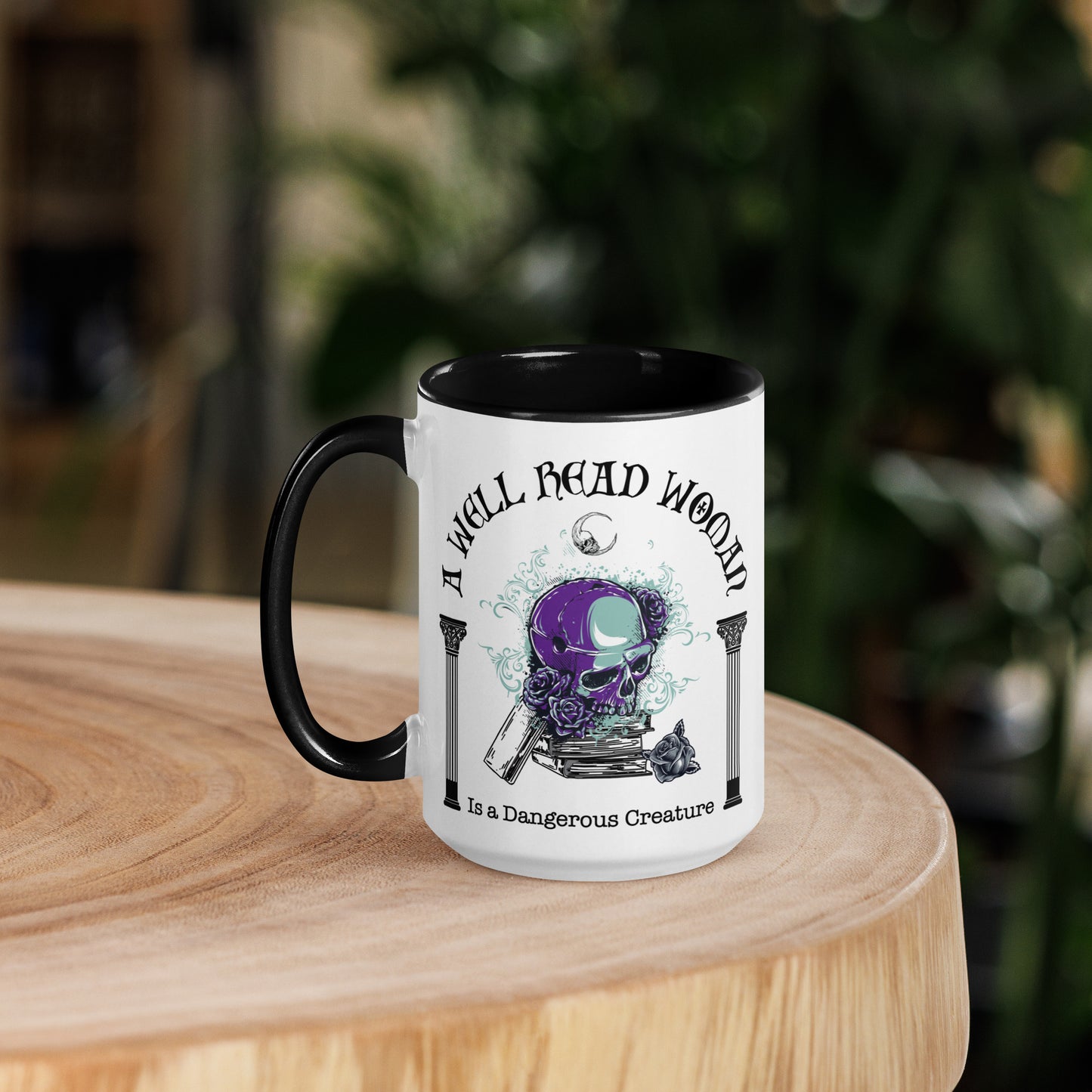 Black and white ceramic coffee mug with words A Well Read Woman Is a Dangerous Creature with a purple skull with roses on top of a stack of books from bookish shop Midnight Gypsy Designs.