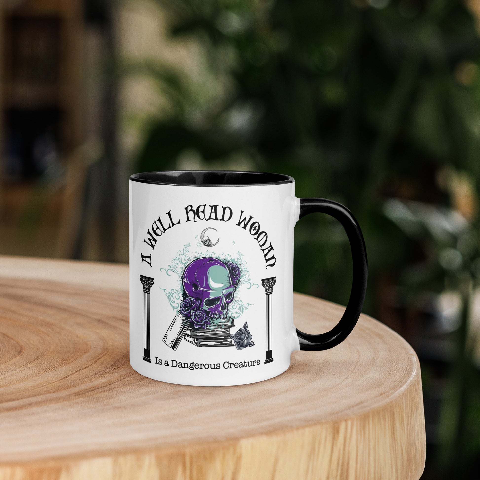 Black and white ceramic coffee mug with words A Well Read Woman Is a Dangerous Creature with a purple skull with roses on top of a stack of books from bookish shop Midnight Gypsy Designs.