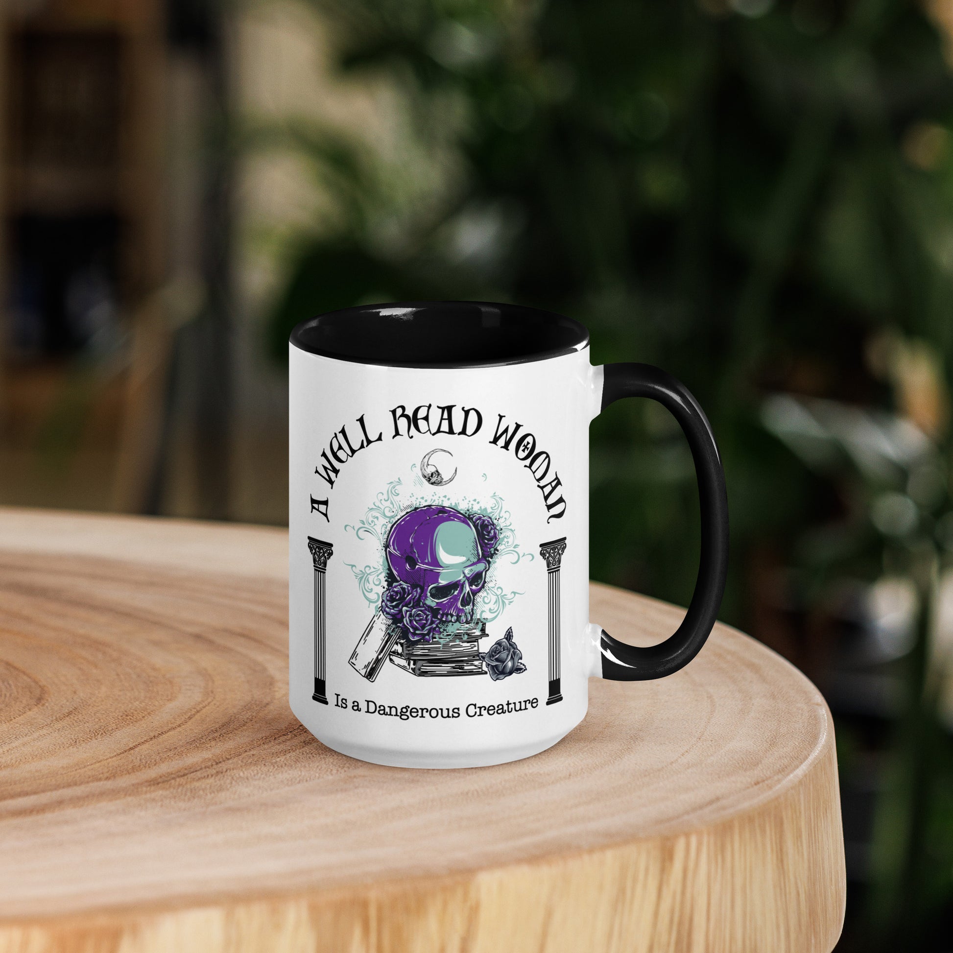 Black and white ceramic coffee mug with words A Well Read Woman Is a Dangerous Creature with a purple skull with roses on top of a stack of books from bookish shop Midnight Gypsy Designs.