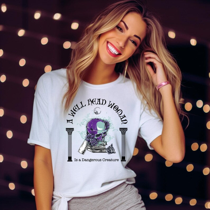 Young woman with long blonde hair smiling wearing a white t-shirt with words A Well Read Woman Is a Dangerous Creature with a purple skull with roses on top of a stack of books from bookish shop Midnight Gypsy Designs.