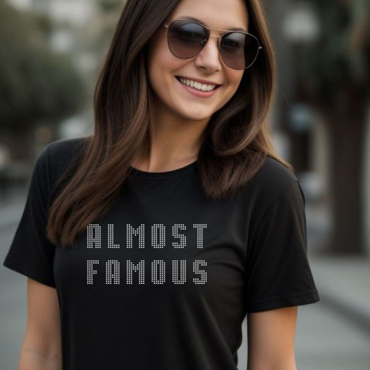 Young woman with long brown hair smiling while standing outside wearing brown tinted sunglasses and a black t-shirt that says Almost Famous with stars created by Midnight Gypsy Designs.