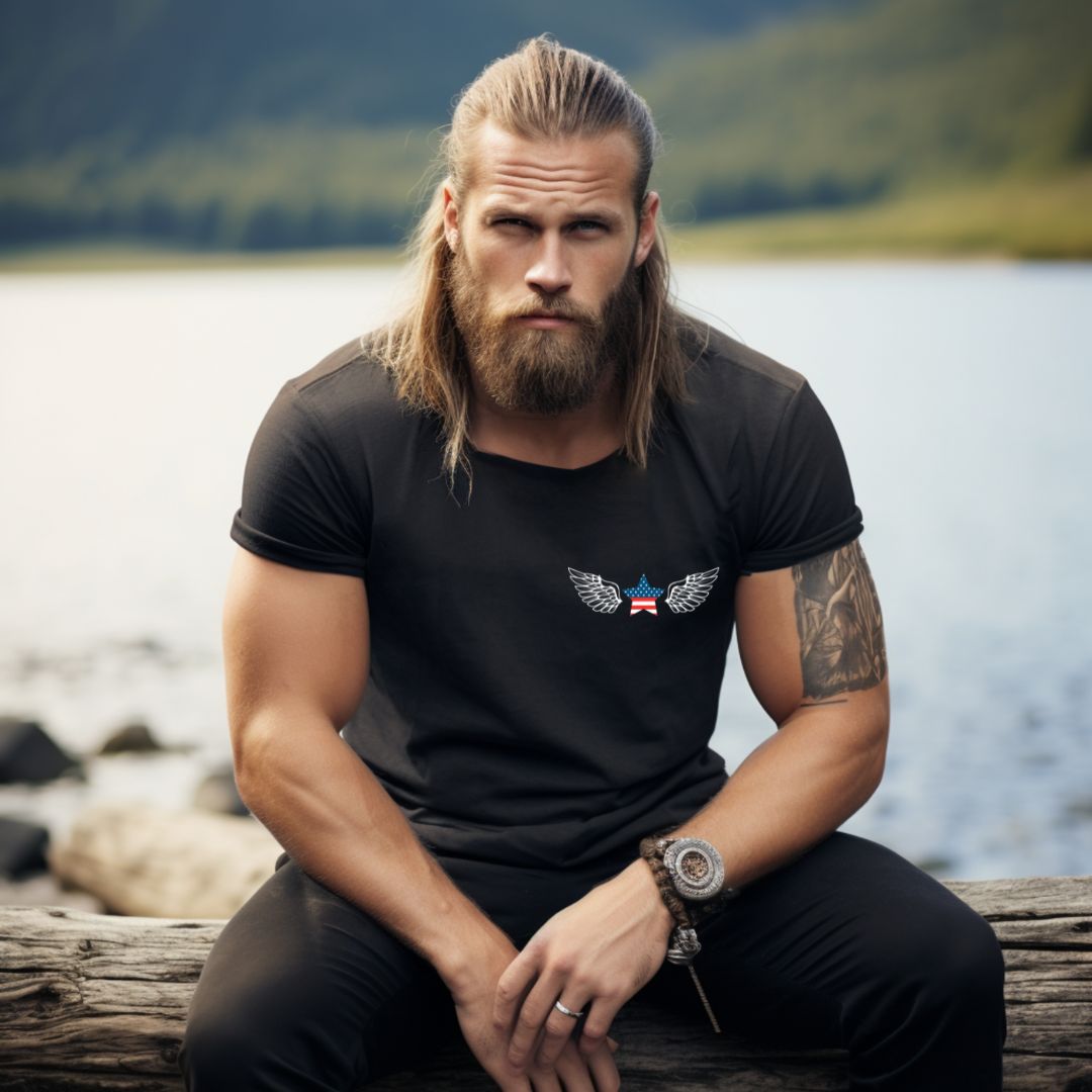 Man with beard and long hair sitting next to a lake wearing a black USA patriotic t-shirt that reads America without her soldiers would be like God without His angles with a USA star with American flag and angel wings to solute American Veterans created by apparel shop MIdnight Gypsy Designs.