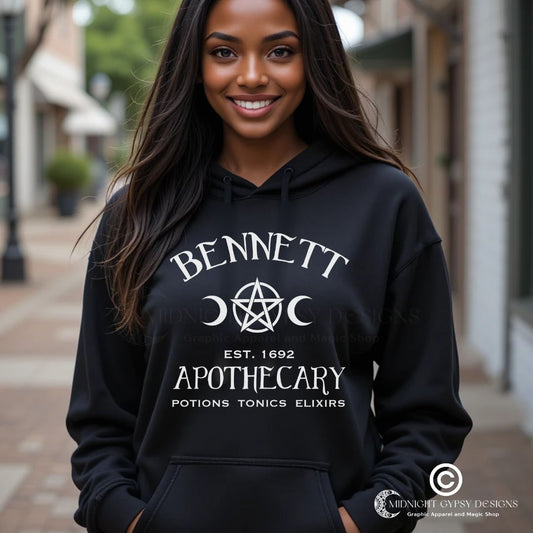 Young woman with long brown hair wearing a Gildan black hoodie with white lettering that reads Bennett Apothecary Established 1692 Potions Tonics Elixirs inspired by Bonnie Bennett from The Vampire Diaries and created by fandom graphic apparel company Midnight Gypsy Designs. Perfect The Vampire Diaries hoodie, Bonnie Bennett hoodie, Bonnie Bennett shirt, Salem witch trials shirt, Mystic Falls hoodie, Vampire Diaries shirt, Mystic Falls shirt, Bonnie Bennett Apothecary shirt, and Salem witch trials hoodie.