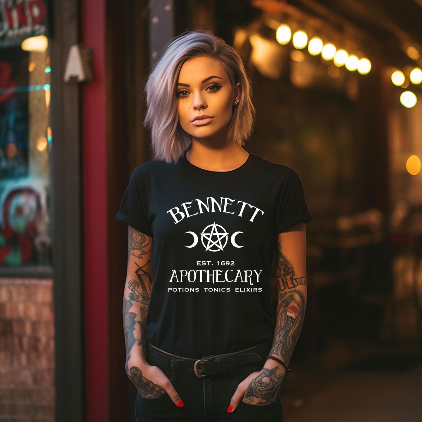 Young woman with shoulder length blonde hair with tattoos on arms and red nail polish with hands in front pockets wearing a black t-shirt with white lettering that reads Bennett Apothecary Established 1692 Potions Tonics Elixirs with witch pentacle and moon inspired by Bonnie Bennett from The Vampire Diaries created by apparel shop Midnight Gypsy Designs.