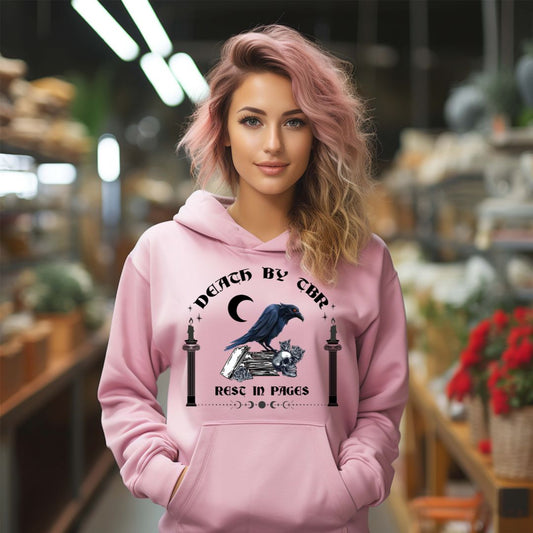 Young woman with long blonde and pink hair standing in a store smiling wearing a pink Gildan hoodie that reads Death By TBR Rest In Pages with moon, raven standing on a book stack, a skull, and two burning candles from bookish shop Midnight Gypsy Designs.