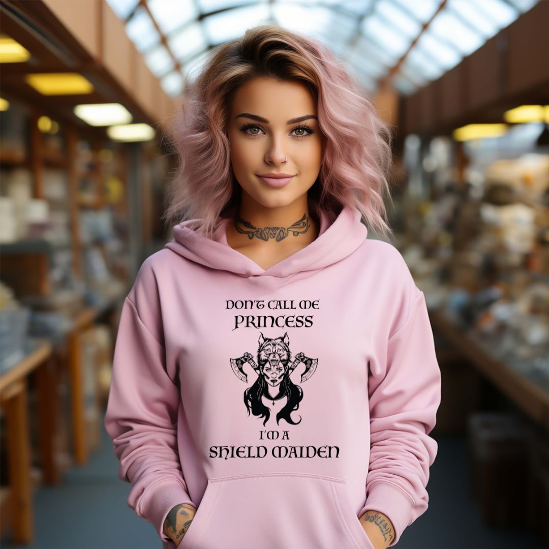 Young woman with pink shoulder length hair smiling while wearing a pink Gildan hoodie with lettering that says Don't Call Me Princess I'm a Shield Maiden with a female Viking Warrior with Norse Viking axes and a wolf created by apparel shop Midnight Gypsy Designs.