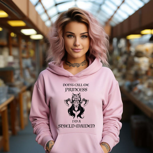 Young woman with pink shoulder length hair smiling while wearing a pink Gildan hoodie with lettering that says Don't Call Me Princess I'm a Shield Maiden with a female Viking Warrior with Norse Viking axes and a wolf created by apparel shop Midnight Gypsy Designs.