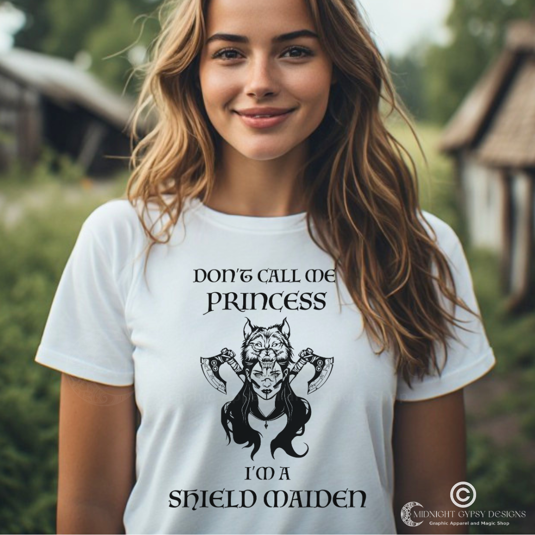 Young Viking woman with long brown hair  wearing a white Shield Maiden Viking Warrior shirt with lettering that says Don't Call Me Princess I'm a Shield Maiden with a female Viking Warrior with Norse Viking axes and Fenrir wolf created by graphic apparel company Midnight Gypsy Designs a part of its Norse Viking Clothing Line. Perfect Viking Shield Maiden shirt and Norse Viking shirt for women.