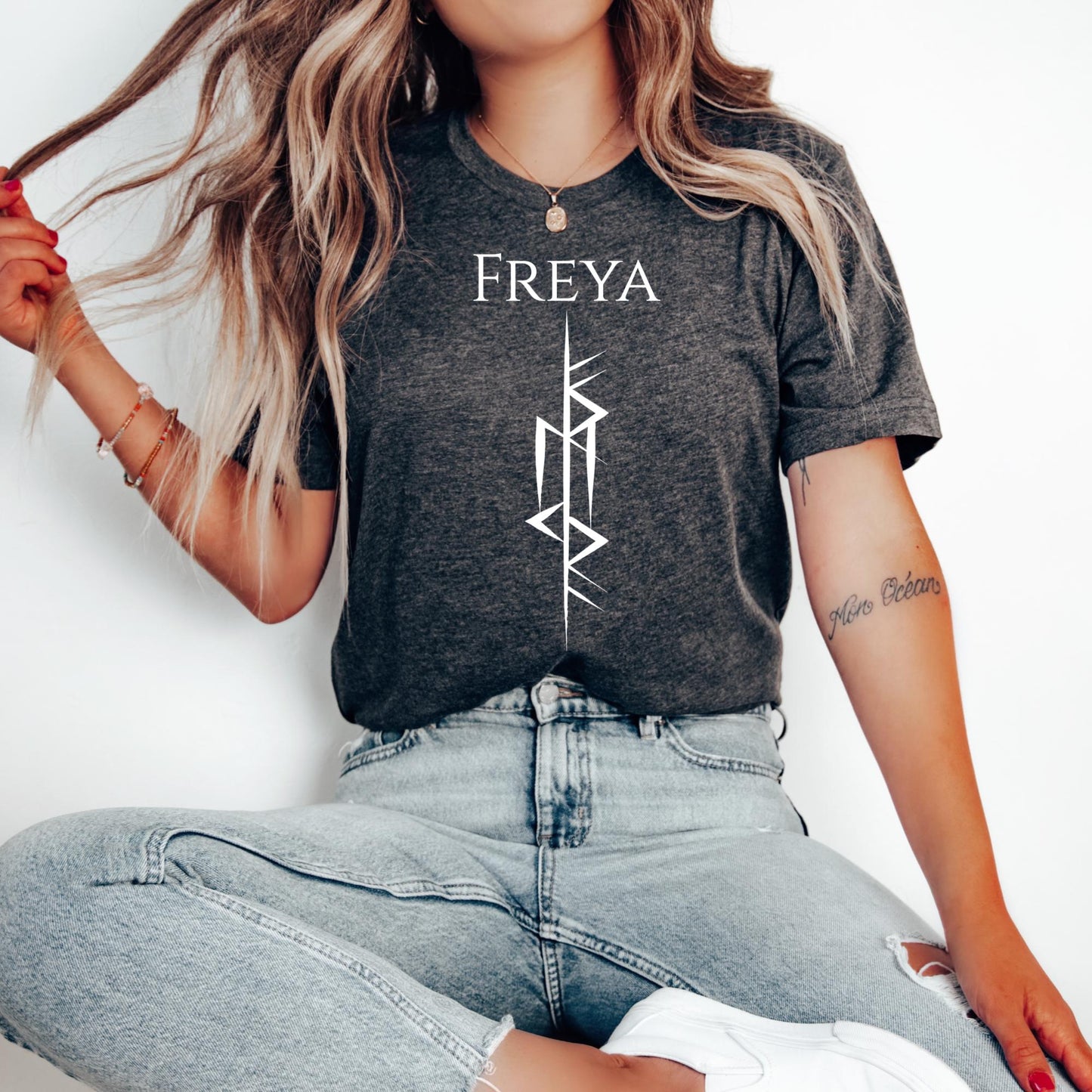 Young woman with long blonde hair with tattoo on arm and red nail polish wearing jeans and a dark grey heather t-shirt that says Freya with the Freya Goddess of Love Norse rune symbol created by Viking apparel shop Midnight Gypsy Designs.