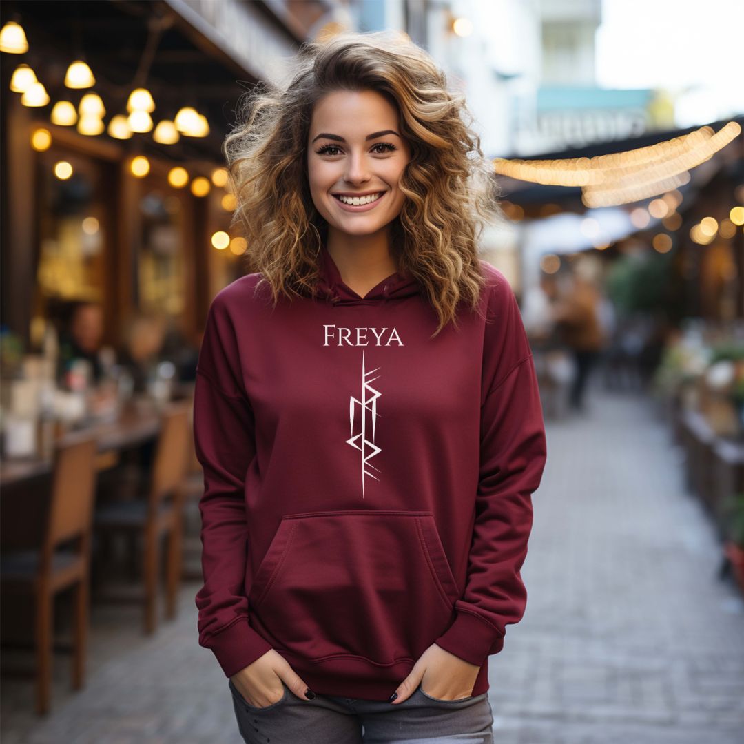 Young woman with blonde wavy hair smiling with hands in her jean pockets standing outside at a restaurant wearing a maroon Viking warrior hoodie that says Freya with the Freya Goddess of Love Norse rune symbol created by Viking apparel shop Midnight Gypsy Designs.