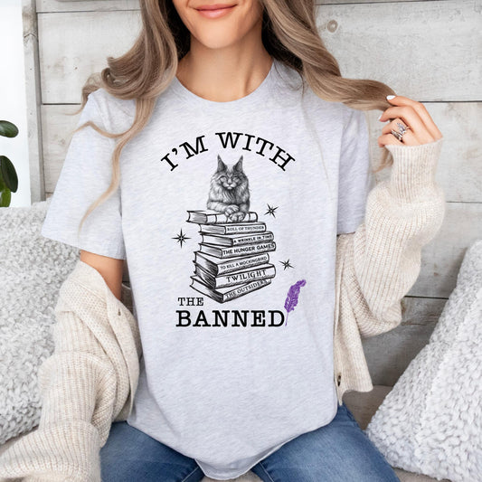 Young woman with long blonde hair wearing a white sweater and a grey t-shirt that reads I'm With the Banned that has a cat sitting on top of a stack of banned books created by Midnight Gypsy Designs.