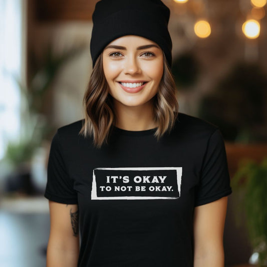 Young woman with shoulder length light brown hair smiling while wearing a black knitted hat and a black kindness t-shirt that reads It's Okay to Not Be Okay created by apparel shop Midnight Gypsy Designs.