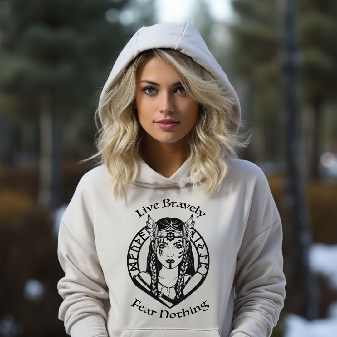 Young woman with wavy blonde hair standing in the woods wearing a Gildan white hoodie that reads Live Bravely Fear Nothing with a Viking Warrior woman with Futhark runes created by apparel shop Midnight Gypsy Designs.