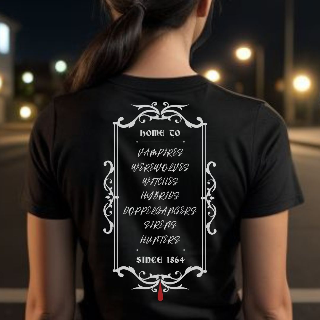 Young woman with long brown pony tail standing outside at night wearing a black Vampire Diaries t-shirt that reads Home to vampires, werewolves, witches, hybrids, doppelgangers, sirens, and hunters since 1864 with a red blood drip created by clothing apparel shop Midnight Gypsy Designs.