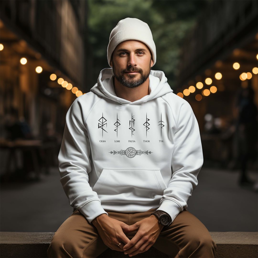 Young man with beard and mustache sitting on bench outside at night wearing a white hat, brown pants, and a Gildan white hoodie that reads Odin, Loki, Freya, Thor, and Tyr with Futhark rune symbols and vegvisir viking compass created by Norse Viking apparel shop Midnight Gypsy Designs. 