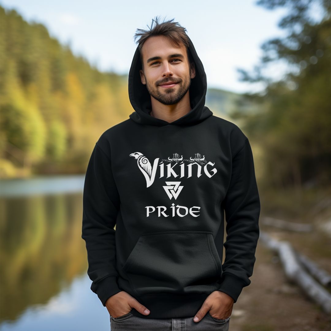 Young man with beard and mustache standing outside by river wearing a Gildan black hoodie that reads Viking Pride with crow and valknut symbol created by Norse Viking apparel shop Midnight Gypsy Designs. 