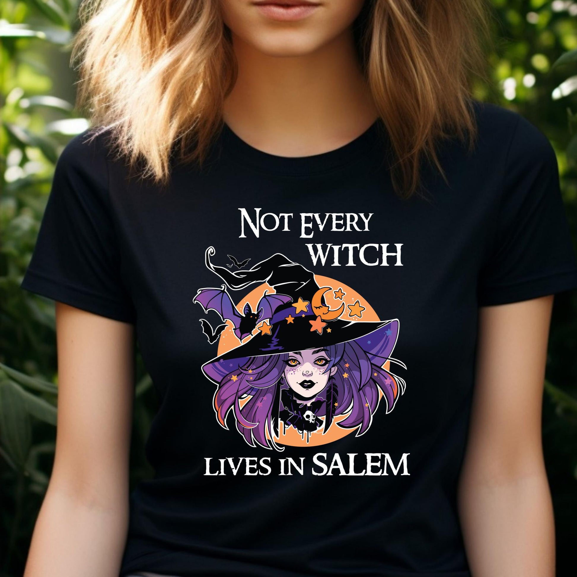 Young woman with shoulder length blonde hair outside in front of green plants wearing a black t-shirt that reads Not Every Witch Lives in Salem with a witch with purple hair wearing a black witch hat in front of a golden moon with stars and black bats created by apparel shop Midnight Gypsy Designs.