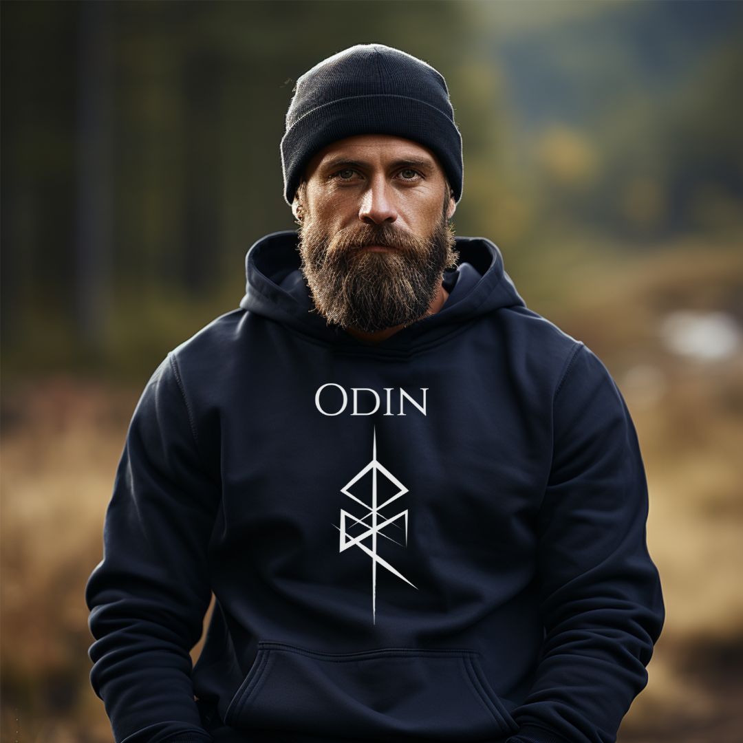 Man with beard and mustache standing outside in woods wearing a navy stocking hat and a Gildan navy hoodie that reads Odin with the Odin God of War Futhark rune symbol created by Norse Viking apparel shop Midnight Gypsy Designs. 