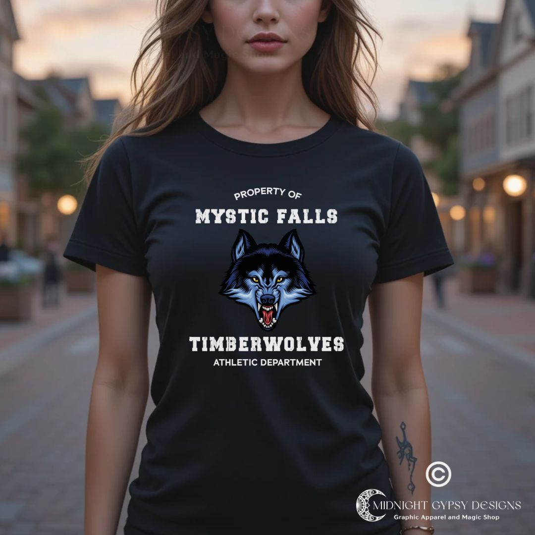 Young woman with long brown hair standing in Mystic Falls wearing a black Mystic Falls Timberwolves shirt with a blue werewolf showing its fangs created by graphic apparel company Midnight Gypsy Designs as part their The Vampire Diaries merch collection. Perfect The Vampire Diaries shirt, Mystic Falls shirt, Mystic Falls Timberwolves t-shirt, Stefan Salvatore shirt, Stefan Salvatore t-shirt, Stefan Salvatore tee, Stefan Salvatore 17 shirt, Mystic Falls High School shirt, Team Stefan shirt, Team Damon Shirt.