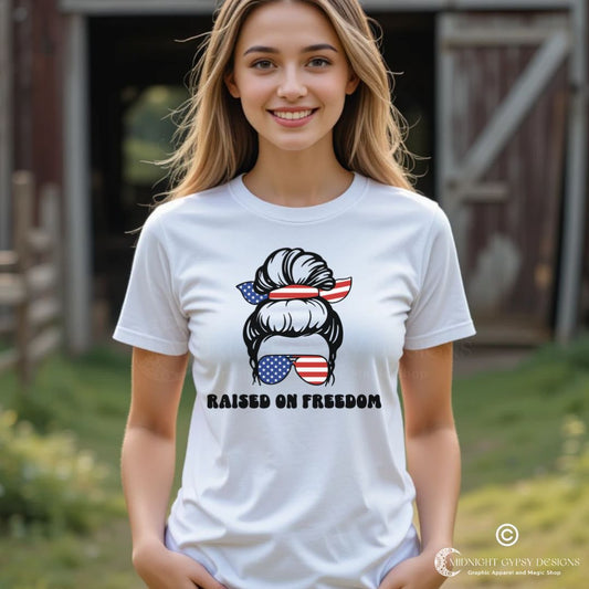 Young woman standing in front of a barn wearing a white Raised on Freedom shirt with graphic of a woman with an American flag hair tie and sunglasses created by graphic apparel company Midnight Gypsy Designs as part of their patriotic apparel collection. Perfect USA patriotic shirt, All American Girl shirt, girl power shirt, American pride shirt, USA shirt, USA flag shirt, womens patriotic shirt, Fourth of July shirt, Patriotic pride shirt, American flag shirt, Independence Day shirt, & American girl shirt.