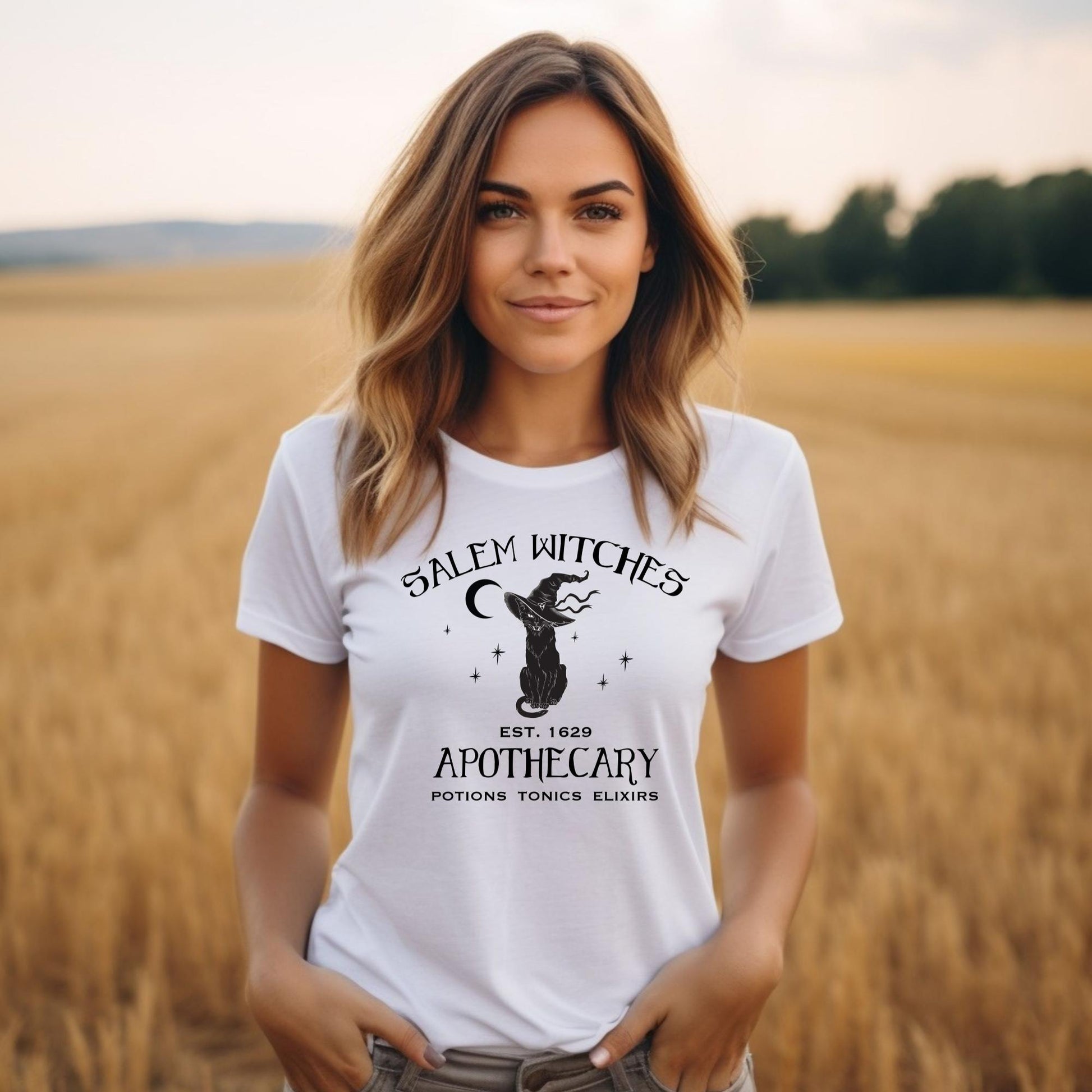 Young woman with long brown hair standing in a wheat field wearing a white t-shirt with the words Salem witches apothecary Est. 1629 with a black cat wearing a witches hat created by clothing apparel shop Midnight Gypsy Designs.