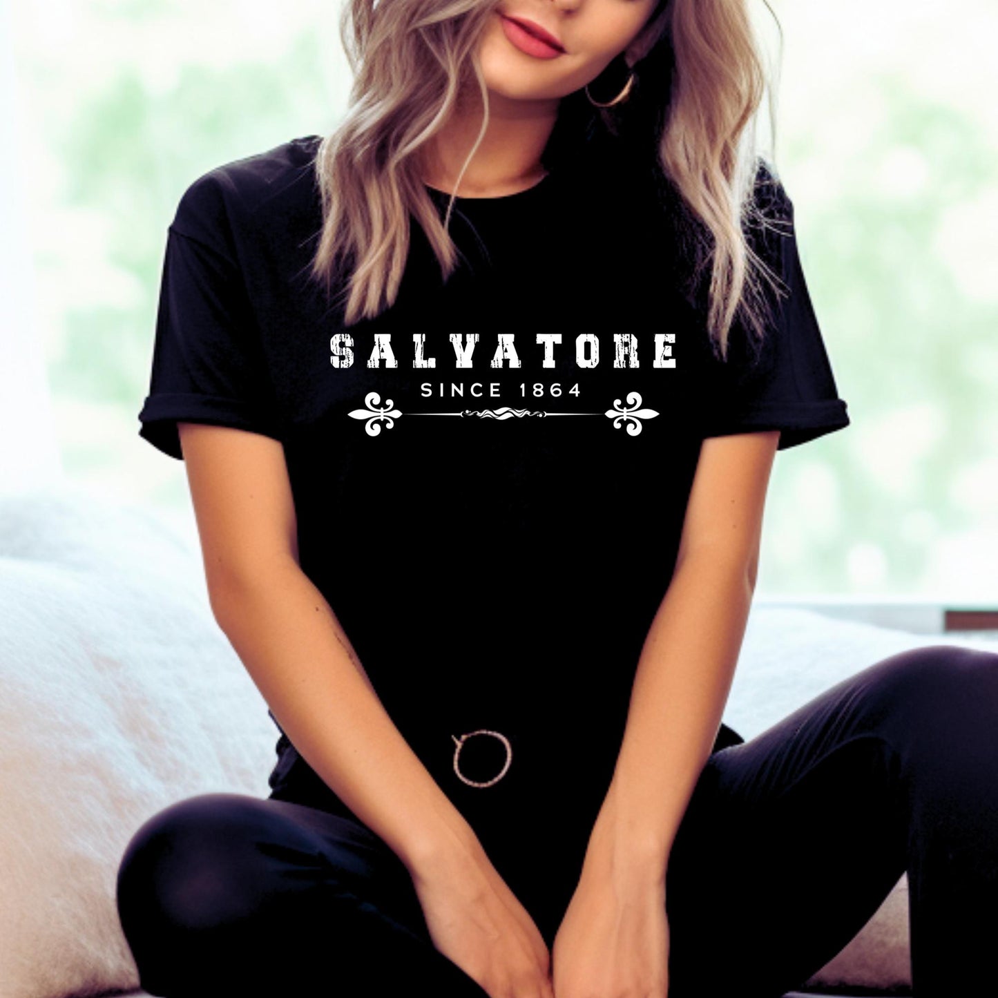 Young woman with blonde hair sitting next to white pillows wearing a black t-shirt that reads Salvatore Since 1864 with fleur de lis created by bookish apparel shop Midnight Gypsy Designs.