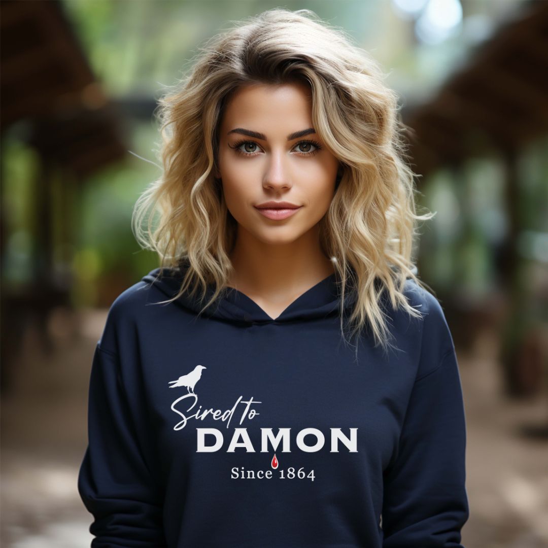 Young woman with wavy blonde hair wearing a Gildan navy The Vampire Diaries Sired to Damon Since 1864 hoodie with a crow and blood drip created by bookish shop Midnight Gypsy Designs.