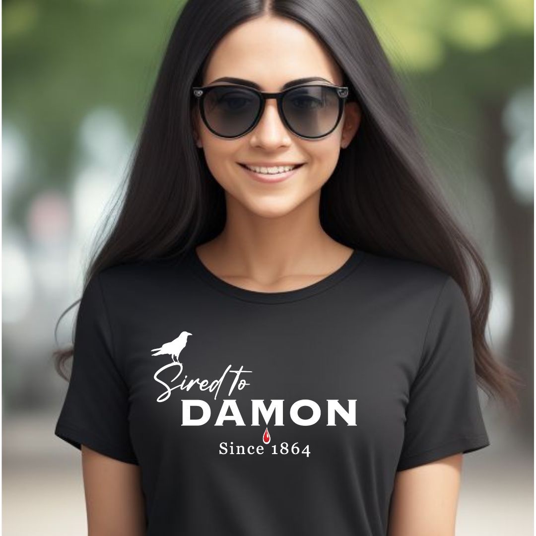 Young woman with long brown hair wearing black sunglasses and a black The Vampire Diaries Sired to Damon Since 1864 t-shirt with a crow and blood drip created by bookish shop Midnight Gypsy Designs.