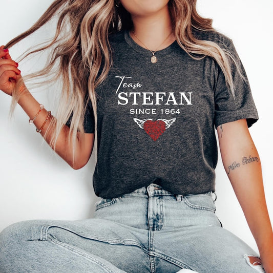 Young woman with long blonde hair wearing denim jeans and a dark grey heather t-shirt that reads The Vampire Diaries Team Stefan Since 1864 with a red heart  created by bookish shop Midnight Gypsy Designs.