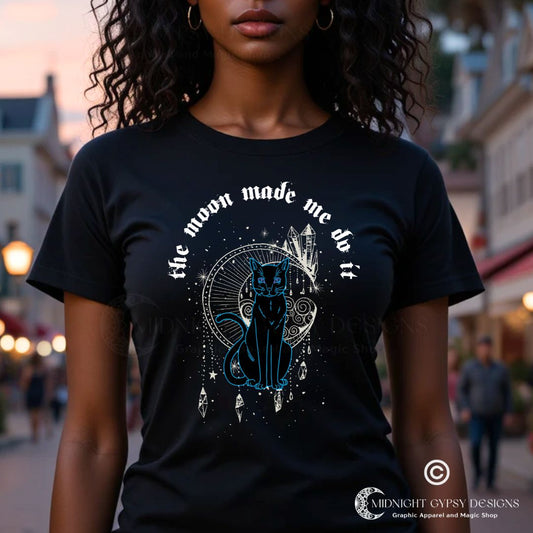 Young woman with long black curly hair and brown eyes smiling with town lights behind her while wearing a black The Moon Made Me Do It t-shirt with a black cat, celestial moon, and crystals created by graphic apparel company Midnight Gypsy Designs as part of their witchy and gothic apparel clothing line Perfect Halloween shirt or cute witchy shirt to wear year round.