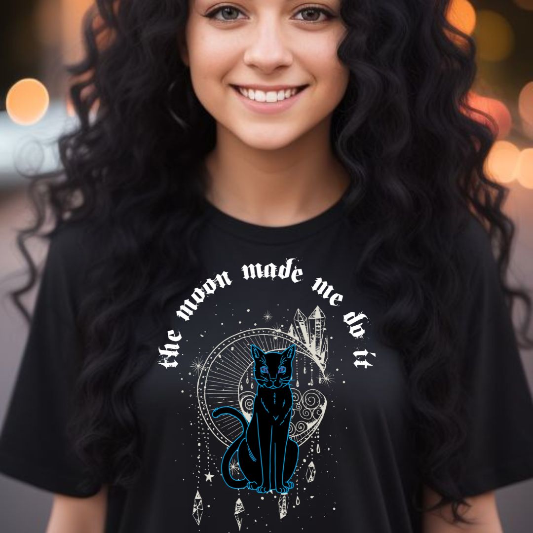 Young woman with long black curly hair and brown eyes smiling with lights behind her while wearing a black t-shirt that reads the moon made me do it with a black cat, celesital moon, and crystals created by clothing apparel shop Midnight Gypsy Designs.