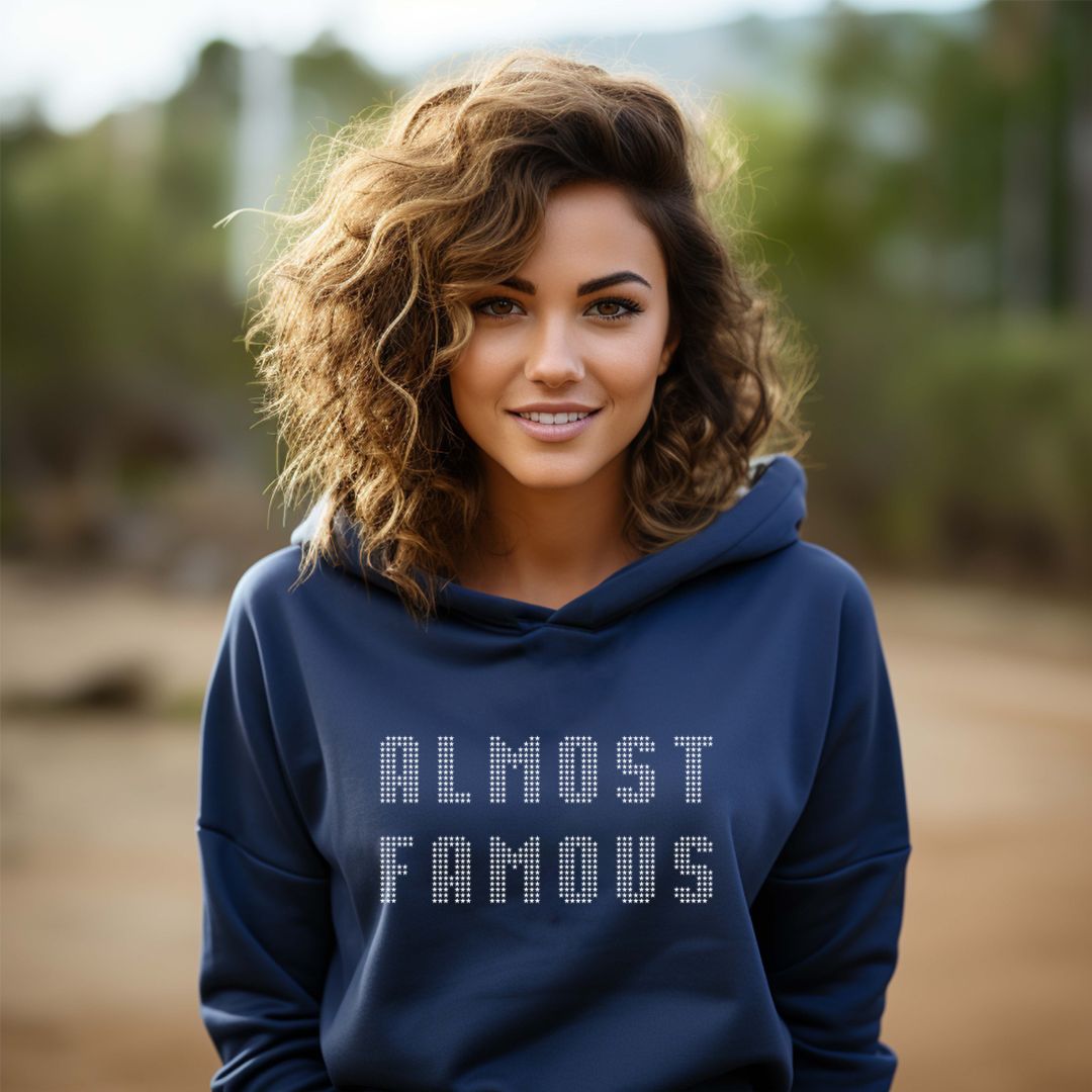 Young woman with shoulder length curly blonde hair standing outside smiling while wearing a Gildan navy hoodie that says Almost Famous created by apparel shop Midnight Gypsy Designs.