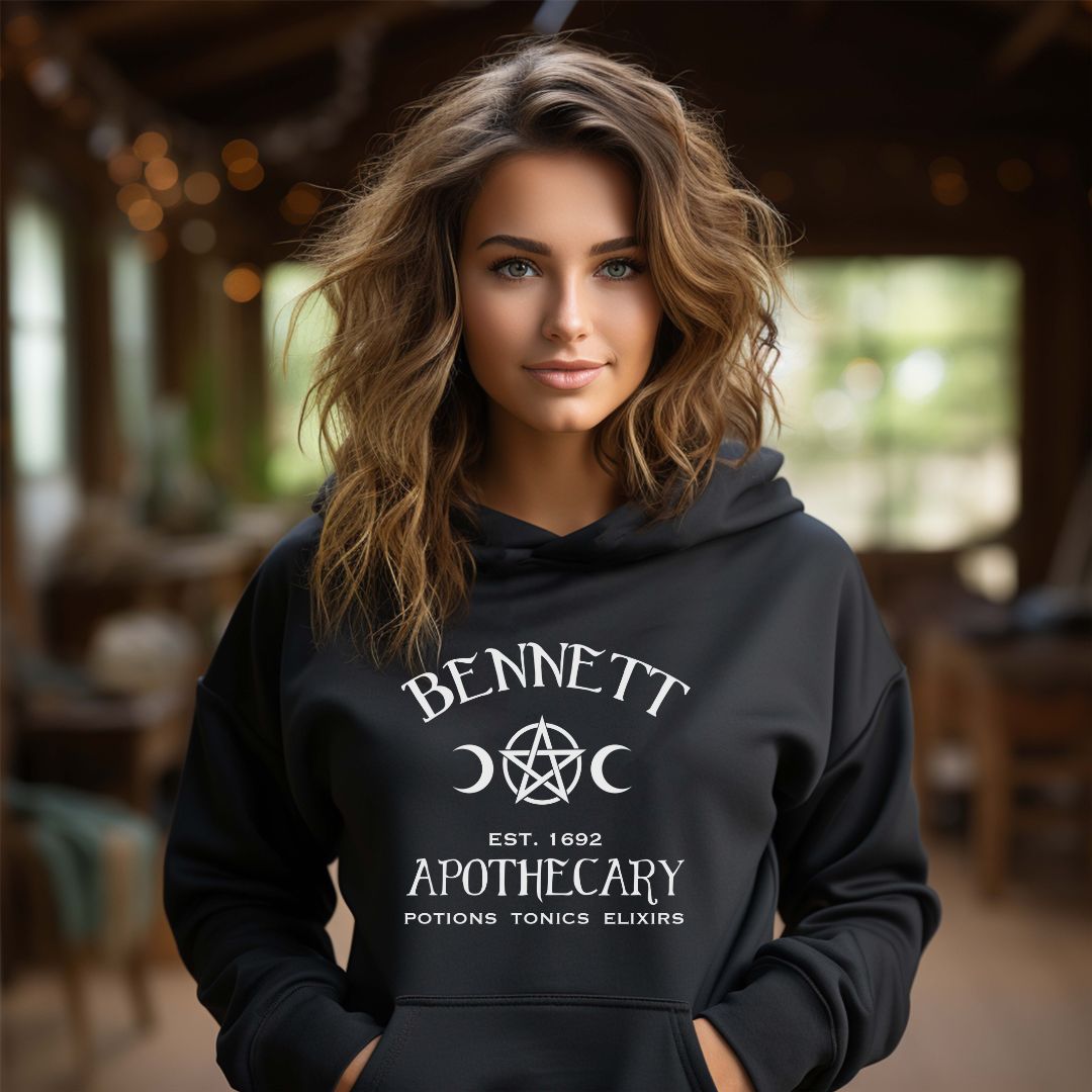 Young woman with long light brown hair wearing a Gildan black hoodie with white lettering that reads Bennett Apothecary Established 1692 Potions Tonics Elixirs inspired by Bonnie Bennett from The Vampire Diaries created by apparel shop Midnight Gypsy Designs.