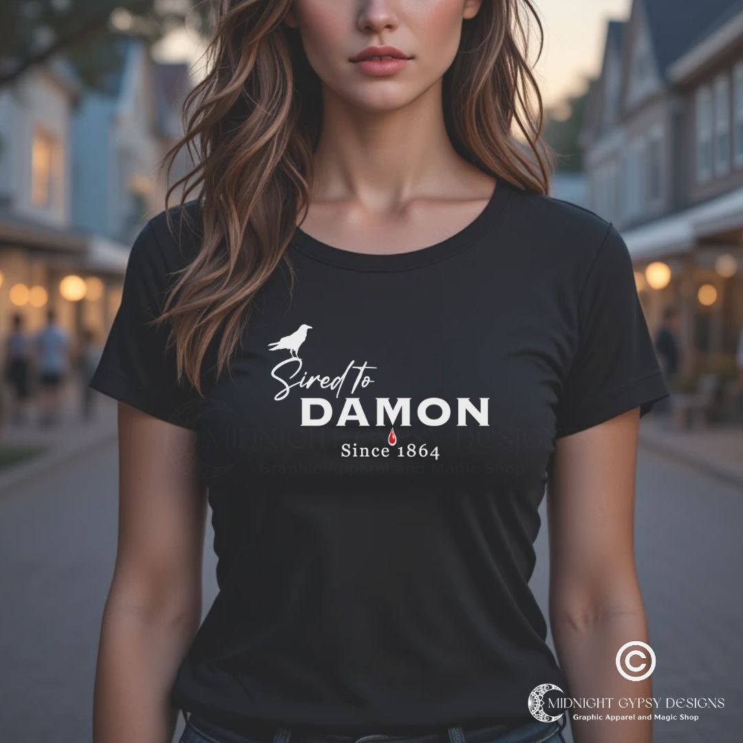 Young woman with long brown hair standing outside in a town square at night wearing a black The Vampire Diaries Sired to Damon Since 1864 t-shirt with a crow and blood drip created by fandom apparel company Midnight Gypsy Designs as part of their The Vampire Diaries clothing line. Perfect Damon Salvatore shirt and Team Damon shirt.