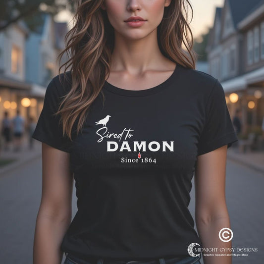 Young woman with long brown hair standing outside in a town square at night wearing a black The Vampire Diaries Sired to Damon Since 1864 t-shirt with a crow and blood drip created by fandom apparel company Midnight Gypsy Designs as part of their The Vampire Diaries clothing line. Perfect Damon Salvatore shirt and Team Damon shirt.
