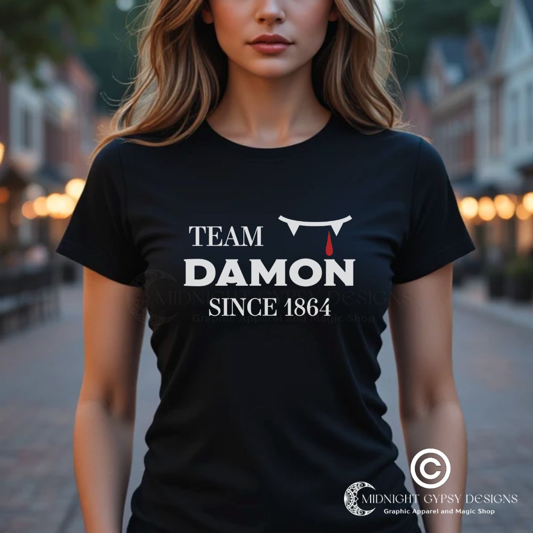 Young woman with long blonde hair wearing a black The Vampire Diaries Team Damon Since 1864 shirt with fangs and blood drip created by graphic apparel company Midnight Gypsy Designs. Perfect   Vampire Diaries Team Damon shirt, Damon Salvatore shirt, the Vampire Diaries shirt, cute vampire shirt, team Damon apparel, Salvatore Brothers shirt, team Damon t-shirt, Mystic Falls shirt, and Mystic Falls t-shirt for fans of The Vampire Diaries!