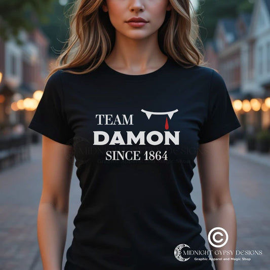 Young woman with long blonde hair wearing a black The Vampire Diaries Team Damon Since 1864 shirt with fangs and blood drip created by graphic apparel company Midnight Gypsy Designs. Perfect   Vampire Diaries Team Damon shirt, Damon Salvatore shirt, the Vampire Diaries shirt, cute vampire shirt, team Damon apparel, Salvatore Brothers shirt, team Damon t-shirt, Mystic Falls shirt, and Mystic Falls t-shirt for fans of The Vampire Diaries!